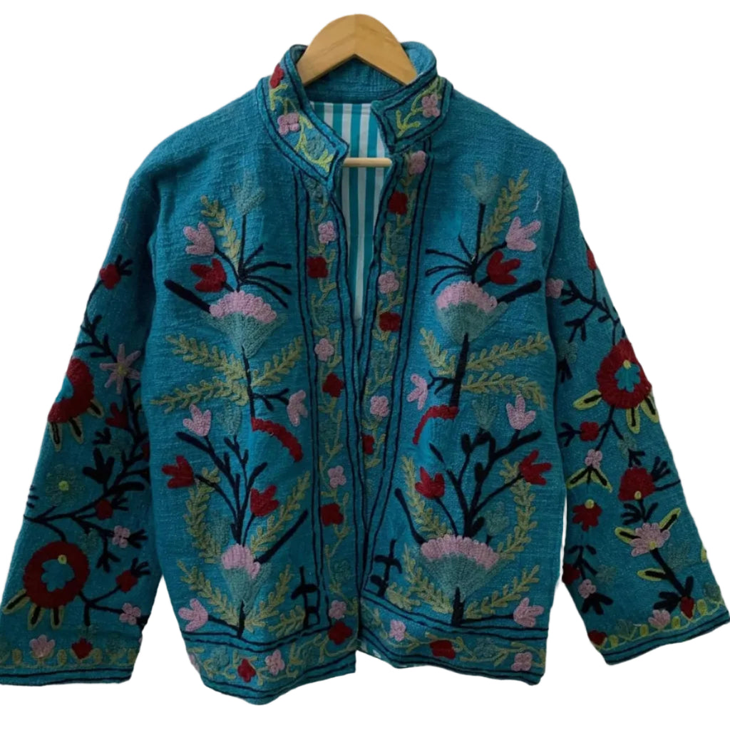 Front view of an open-style handmade teal blue textured cotton TNT short jacket featuring soft pink and deep red Suzani floral embroidery, showcasing a matching teal-and-white striped interior lining, hanging on a wooden hanger against a white wall.