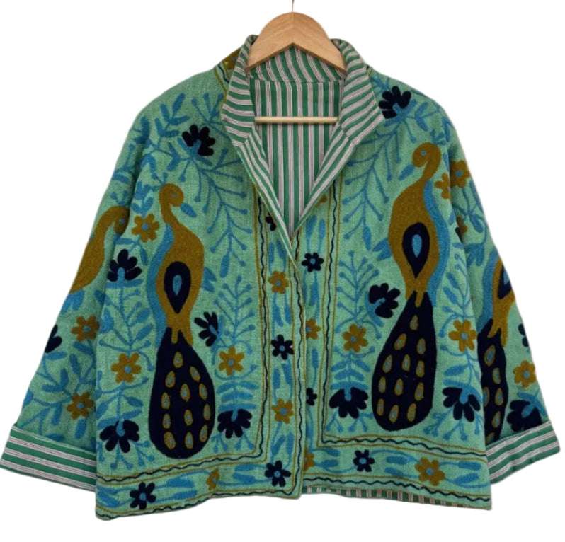 Front view of an open-style handmade mint green textured cotton TNT jacket featuring Suzani embroidery of two majestic peacocks in gold and navy blue, showcasing a green-and-white striped interior lining on the collar and folded cuffs, isolated on a solid wh