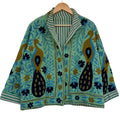 Front view of an open-style handmade mint green textured cotton TNT jacket featuring Suzani embroidery of two majestic peacocks in gold and navy blue, showcasing a green-and-white striped interior lining on the collar and folded cuffs, isolated on a solid wh
