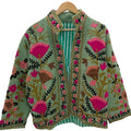 Front close-up of an open-style handmade mint green textured cotton TNT jacket featuring vibrant pinkish-red pomegranate and orange floral Suzani embroidery with thick dark green vines, hanging on a wooden hanger against a white wall.