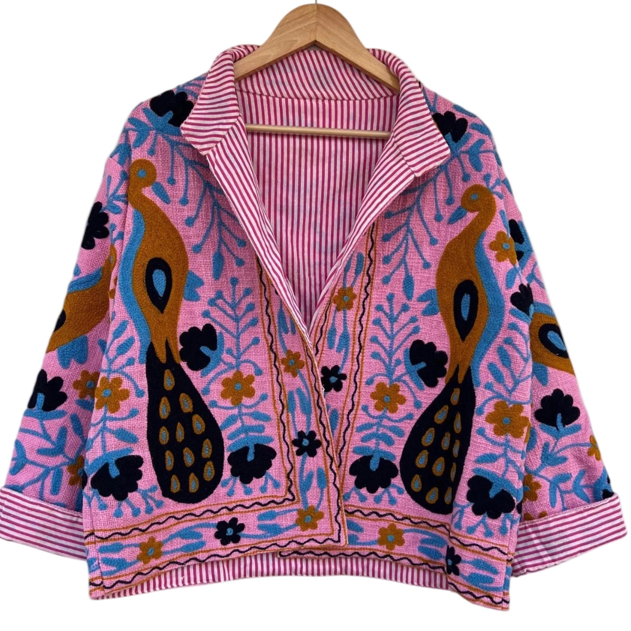Front view of an open-style handmade bright pink textured cotton TNT jacket featuring Suzani embroidery of two majestic peacocks in gold and navy blue, showcasing a pink-and-white striped interior lining on the collar and folded cuffs, isolated on a solid white background.