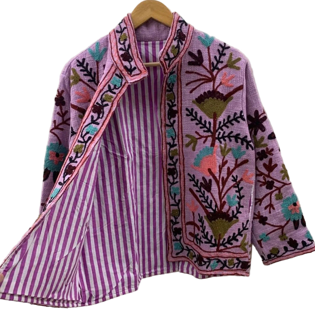 Front view of a handmade lilac textured cotton TNT short jacket with its lapel held open to reveal a striking purple-and-white striped inner lining. The exterior features vibrant teal, coral pink, and olive green Suzani floral embroidery.