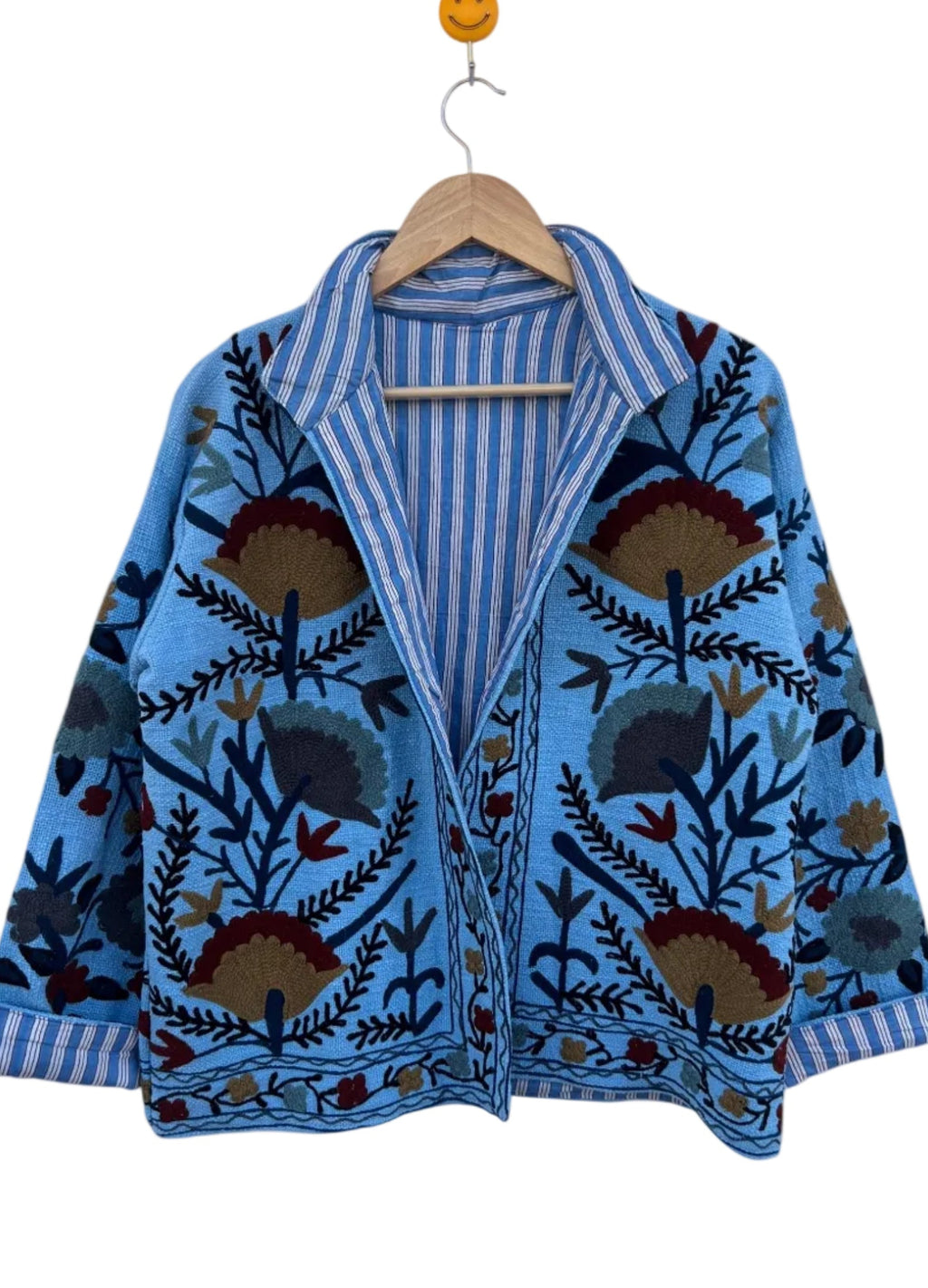 Angled front view of an open-style handmade light blue textured cotton TNT jacket featuring Suzani floral embroidery in dark red, gold, and navy, showcasing a blue-and-white striped interior lining on the collar, isolated on a solid white background.