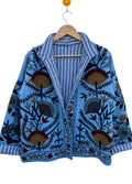 Angled front view of an open-style handmade light blue textured cotton TNT jacket featuring Suzani floral embroidery in dark red, gold, and navy, showcasing a blue-and-white striped interior lining on the collar, isolated on a solid white background.
