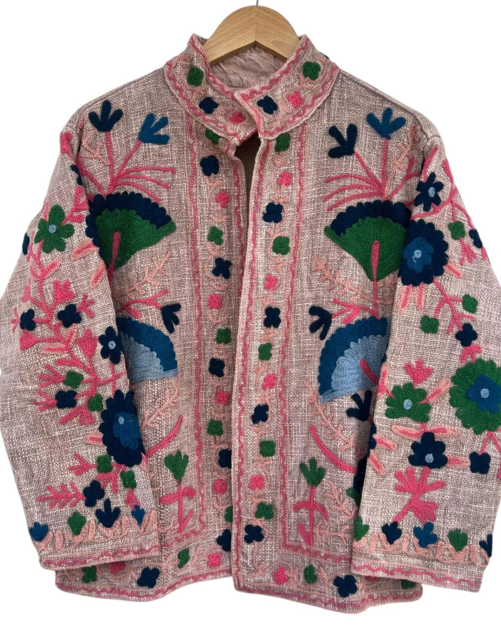 Light pink textured jacket featuring intricate, vibrant floral embroidery in hot pink, green, navy, and light blue, hanging on a wooden hanger.