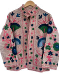 Light pink textured jacket featuring intricate, vibrant floral embroidery in hot pink, green, navy, and light blue, hanging on a wooden hanger.