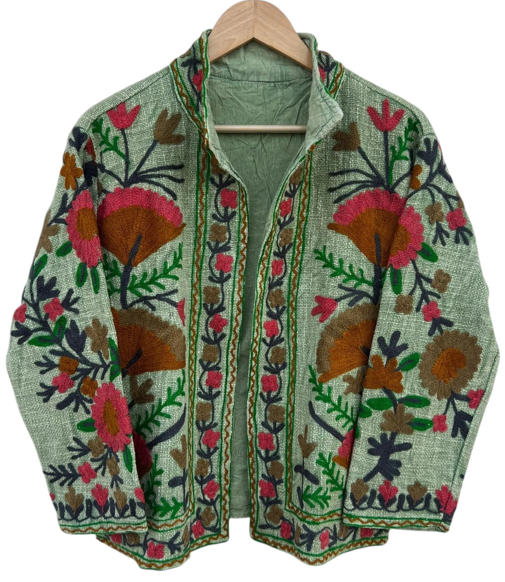 Front view of an open-style handmade sage green textured cotton TNT short jacket featuring vibrant pink and rust Suzani floral embroidery with dark green vines, isolated on a solid white background.