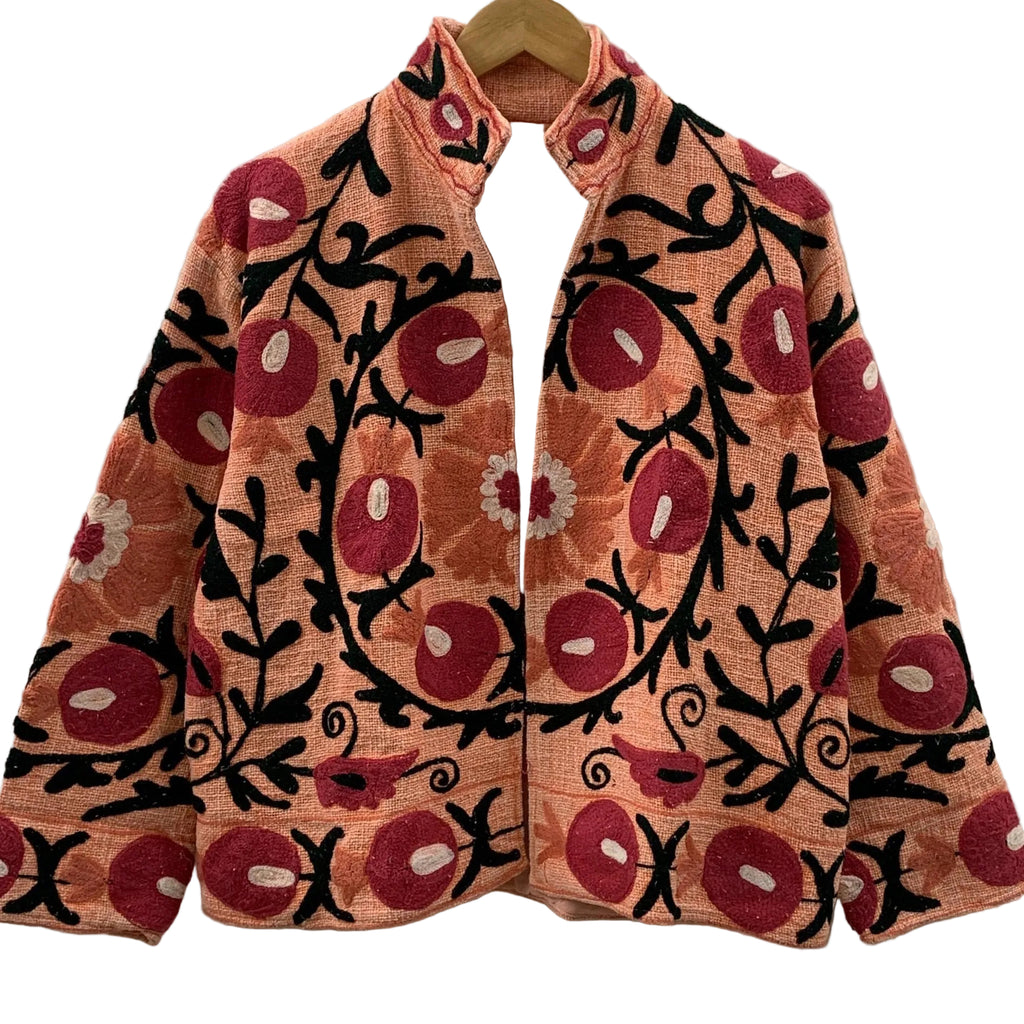 Front view of an open-style handmade peach textured cotton TNT short jacket featuring vibrant red pomegranate and orange floral Suzani embroidery with thick black vines, hanging on a wooden hanger against a white wall.