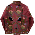 Back view of a handmade maroon textured cotton TNT short jacket featuring intricate Suzani embroidery with olive green vines, pink floral fans, and black botanical accents, hanging on a wooden hanger against a white wall.