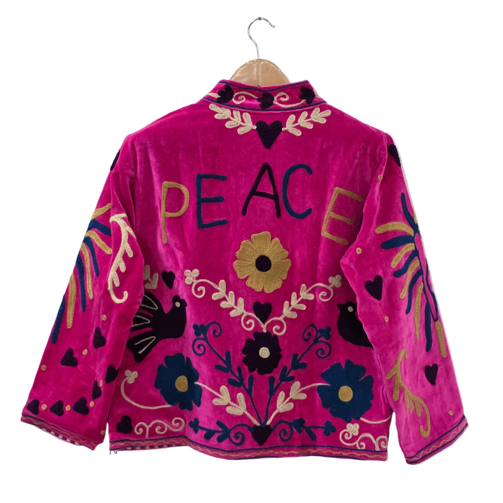 A bright fuchsia pink velvet bohemian jacket shown from the back on a wooden hanger. The word PEACE is embroidered in large blue and gold letters across the upper back. The traditional Suzani embroidery design also includes black silhouette birds, yellow and blue flowers, winding gold vines, and dark heart shapes scattered throughout.