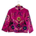 A bright fuchsia pink velvet bohemian jacket shown from the back on a wooden hanger. The word PEACE is embroidered in large blue and gold letters across the upper back. The traditional Suzani embroidery design also includes black silhouette birds, yellow and blue flowers, winding gold vines, and dark heart shapes scattered throughout.