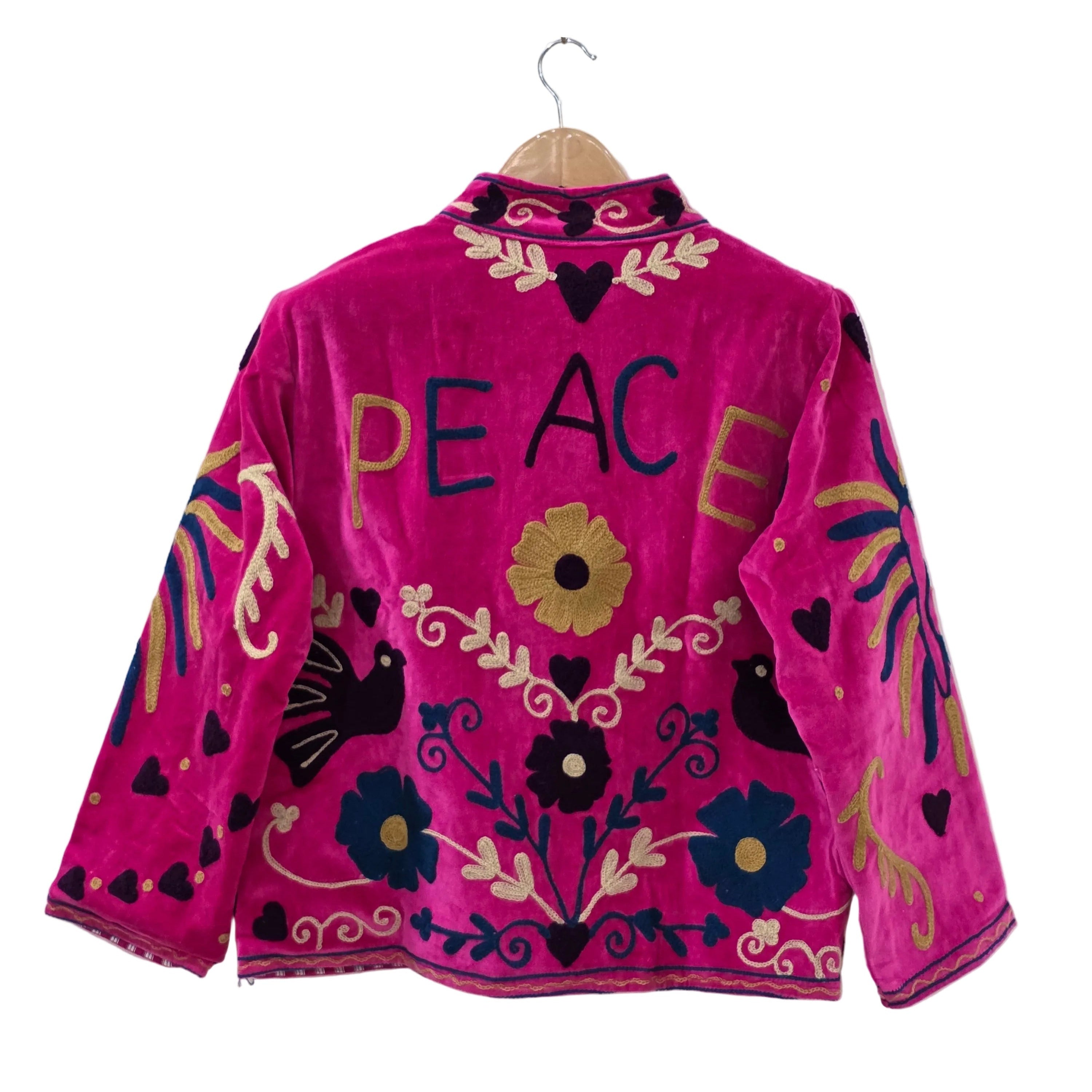 A bright fuchsia pink velvet bohemian jacket shown from the back on a wooden hanger. The word PEACE is embroidered in large blue and gold letters across the upper back. The traditional Suzani embroidery design also includes black silhouette birds, yellow and blue flowers, winding gold vines, and dark heart shapes scattered throughout.