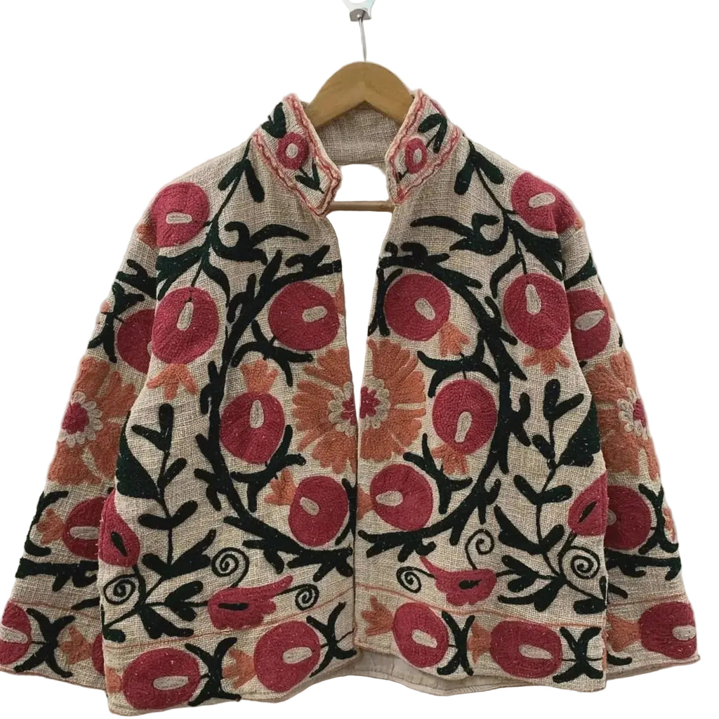 Front view of an open-style handmade beige textured cotton TNT short jacket featuring vibrant red pomegranate and orange floral Suzani embroidery with thick black vines, hanging on a wooden hanger against a white wall.