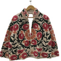 Front view of an open-style handmade beige textured cotton TNT short jacket featuring vibrant red pomegranate and orange floral Suzani embroidery with thick black vines, hanging on a wooden hanger against a white wall.