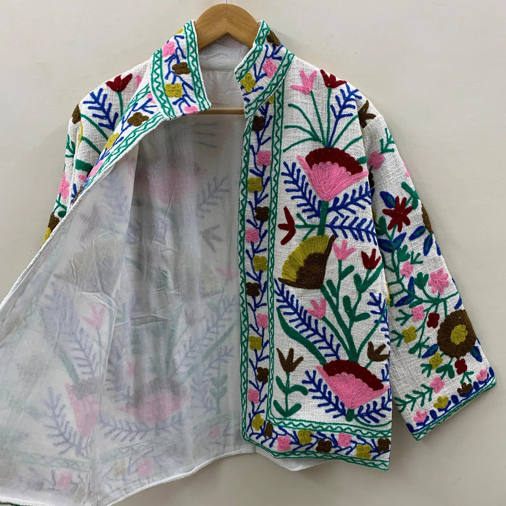 Front view of an open-style handmade white textured cotton TNT short jacket featuring vibrant pink, royal blue, and emerald green Suzani floral embroidery. The left lapel is pulled open to reveal a solid white inner lining, and the jacket is hanging on a wooden hanger against a white wall.