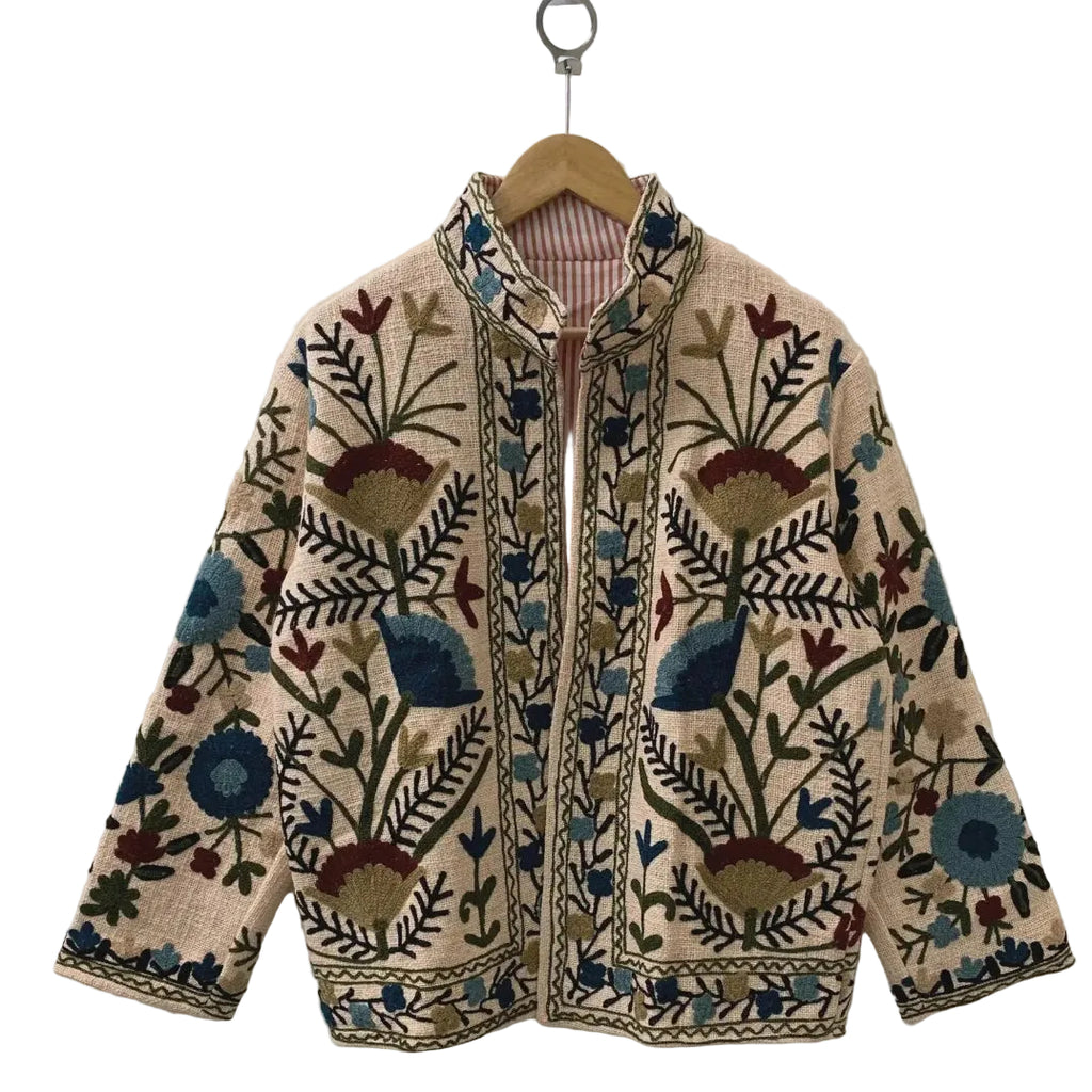 Front view of a handmade beige textured cotton TNT short jacket featuring deep blue, teal, and burgundy Suzani floral embroidery, with a subtle reddish-orange and white striped inner lining visible at the collar, hanging on a wooden hanger against a white wall.