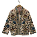 Front view of a handmade beige textured cotton TNT short jacket featuring deep blue, teal, and burgundy Suzani floral embroidery, with a subtle reddish-orange and white striped inner lining visible at the collar, hanging on a wooden hanger against a white wall.