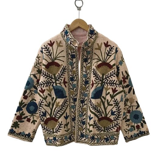 Front view of a handmade beige textured cotton TNT short jacket featuring deep blue, teal, and burgundy Suzani floral embroidery, with a subtle reddish-orange and white striped inner lining visible at the collar, hanging on a wooden hanger against a white wall.