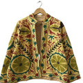 Front view of an open-style handmade yellow textured cotton TNT short jacket featuring large green circular floral Suzani embroidery, earthy brown leaves, and maroon vines. The jacket is slightly open, revealing a yellow-and-white striped interior lining, hanging on a wooden hanger against a white wall.