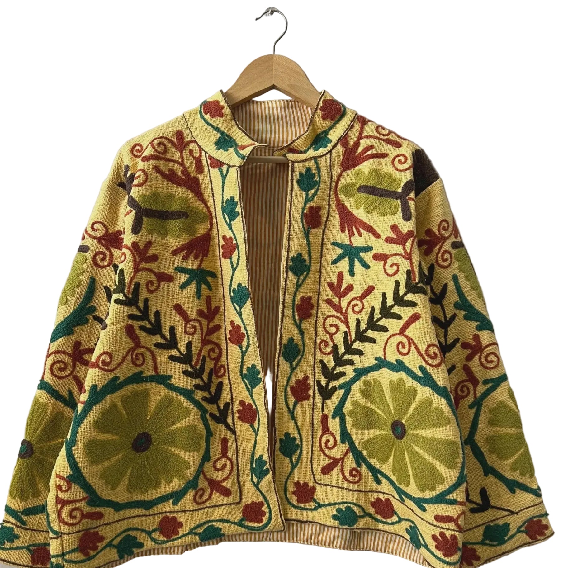 Front view of an open-style handmade yellow textured cotton TNT short jacket featuring large green circular floral Suzani embroidery, earthy brown leaves, and maroon vines. The jacket is slightly open, revealing a yellow-and-white striped interior lining, hanging on a wooden hanger against a white wall.