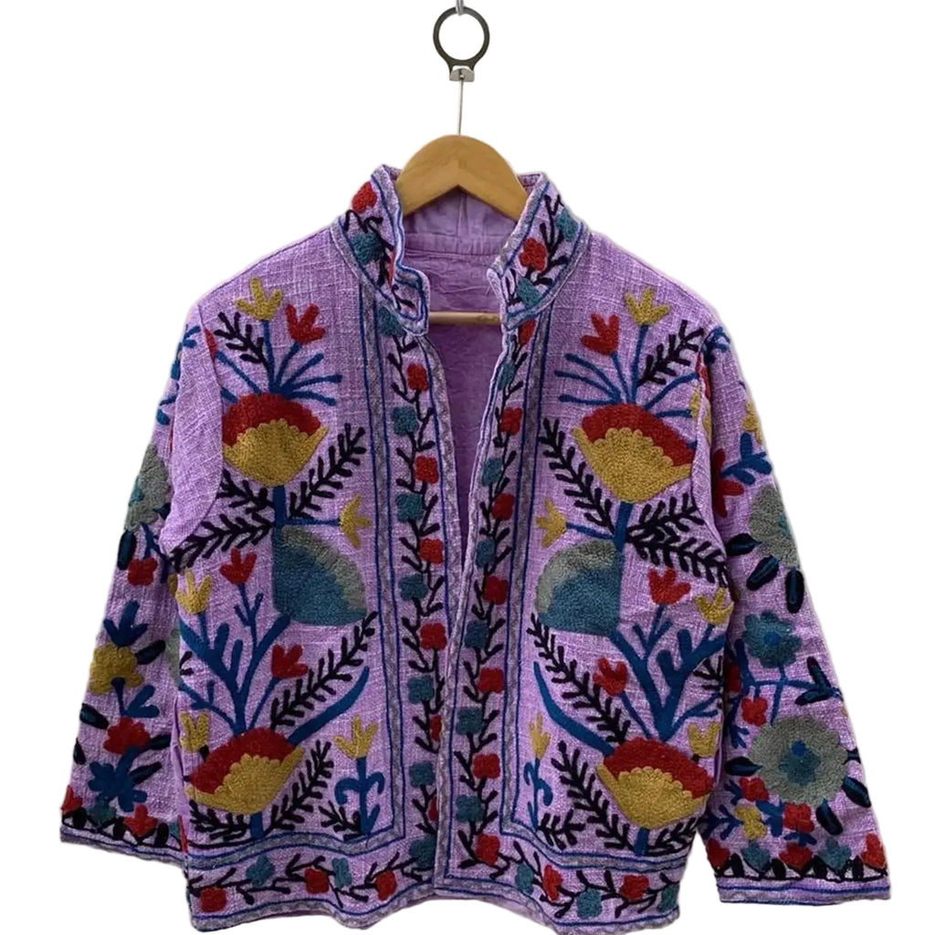 Front view of an open-style handmade lavender textured cotton TNT short jacket featuring mustard yellow, red, and blue Suzani floral embroidery, showcasing a matching solid light purple interior lining, hanging on a wooden hanger against a white wall.