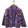 Front view of an open-style handmade lavender textured cotton TNT short jacket featuring mustard yellow, red, and blue Suzani floral embroidery, showcasing a matching solid light purple interior lining, hanging on a wooden hanger against a white wall.