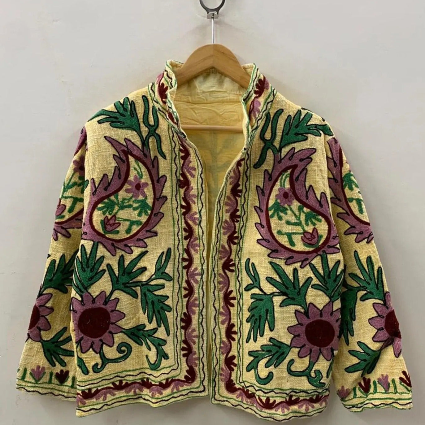 Front view of an open-style handmade pale yellow textured cotton TNT short jacket featuring large mauve paisley motifs, green leaves, and maroon Suzani floral embroidery, showcasing a matching solid yellow interior lining, hanging on a wooden hanger against a white wall.