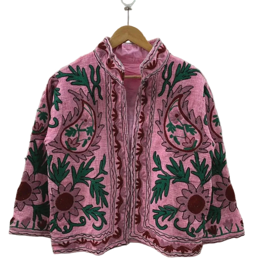Front view of an open-style handmade pink textured cotton TNT short jacket featuring large maroon paisley motifs, deep green leaves, and Suzani floral embroidery, showcasing a matching solid pink interior lining, hanging on a wooden hanger against a white wall.