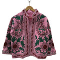 Front view of an open-style handmade pink textured cotton TNT short jacket featuring large maroon paisley motifs, deep green leaves, and Suzani floral embroidery, showcasing a matching solid pink interior lining, hanging on a wooden hanger against a white wall.