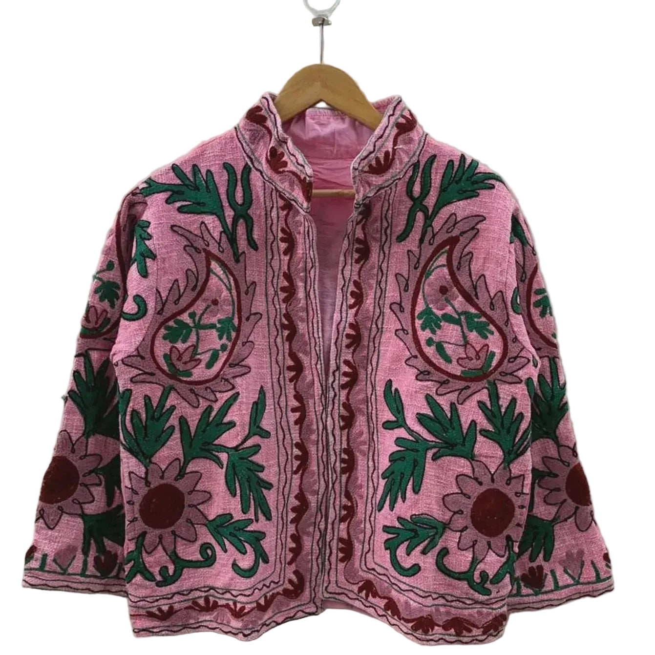 Front view of an open-style handmade pink textured cotton TNT short jacket featuring large maroon paisley motifs, deep green leaves, and Suzani floral embroidery, showcasing a matching solid pink interior lining, hanging on a wooden hanger against a white wall.