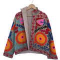 Front view of an open-style handmade khaki textured cotton TNT short jacket featuring vibrant neon orange sunbursts, hot pink flowers, and bright blue Suzani embroidery. The lapel is folded back to reveal a brown-and-white striped interior lining, hanging on a wooden hanger against a white wall.