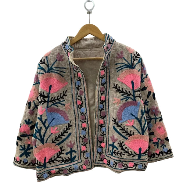 Front view of an open-style handmade beige textured cotton TNT short jacket featuring bright pink, periwinkle blue, and mauve Suzani floral embroidery with dark teal vines, showcasing a matching solid beige interior lining.