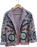 A light purple bohemian jacket on a wooden hanger against a white background. It features Suzani-style embroidery with large teal circular flowers, dark green leaves, black swirls, and an orange embroidered border along the bottom hem. The jacket is open, showing a solid lavender lining.