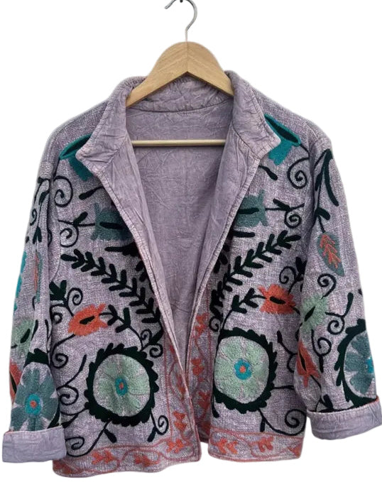 A light purple bohemian jacket on a wooden hanger against a white background. It features Suzani-style embroidery with large teal circular flowers, dark green leaves, black swirls, and an orange embroidered border along the bottom hem. The jacket is open, showing a solid lavender lining.