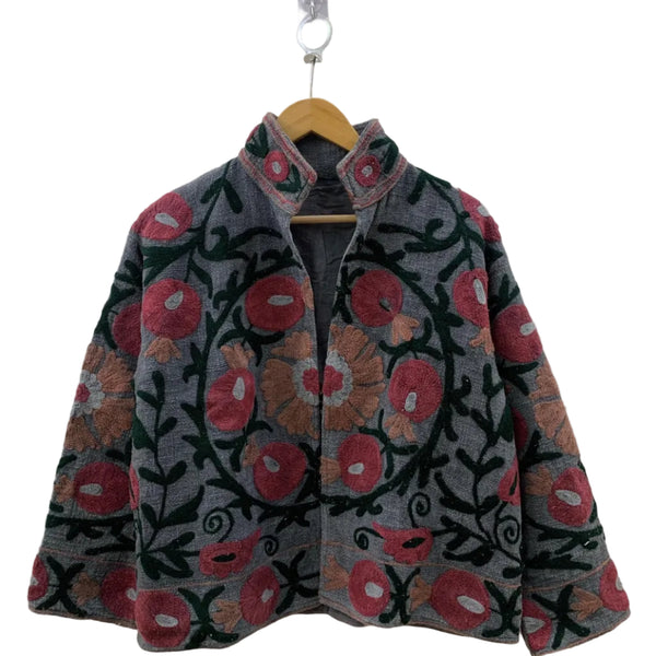 Front view of an open-style handmade dark grey textured cotton TNT short jacket featuring red pomegranate motifs, tan flowers, and dark green Suzani vine embroidery, showcasing a matching solid dark grey interior lining, hanging on a wooden hanger against a white wall .