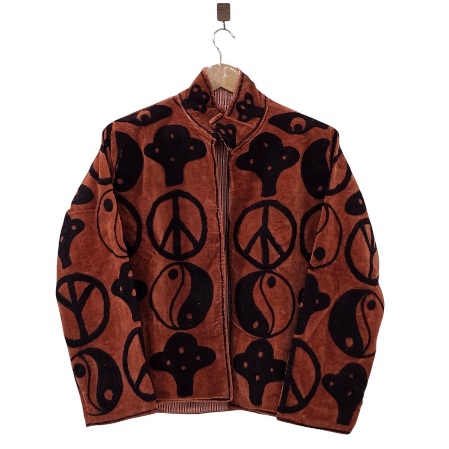 A rust copper velvet statement jacket on a wooden hanger. It features large, contrasting black retro symbols all over, including peace signs, yin-yang symbols, and abstract mystic shapes. The jacket has a stand collar, long sleeves, and an open front showing a peek of striped lining.