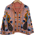Front view of an open-style handmade peachy-pink textured cotton TNT short jacket featuring large mustard yellow and navy blue peacock motifs, light blue vines, and dark floral Suzani embroidery. The collar reveals an orange-and-white striped interior lining, hanging on a wooden hanger against a white wall as seen