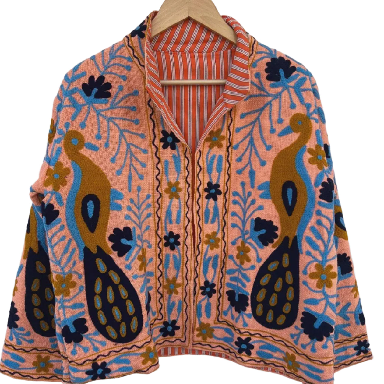 Front view of an open-style handmade peachy-pink textured cotton TNT short jacket featuring large mustard yellow and navy blue peacock motifs, light blue vines, and dark floral Suzani embroidery. The collar reveals an orange-and-white striped interior lining, hanging on a wooden hanger against a white wall as seen
