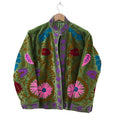 An olive green velvet bohemian jacket on a wooden hanger. It features traditional Suzani embroidery with large pink daisy-like flowers near the hem, bright blue and red leaves, winding bronze vines, and a contrasting purple embroidered border along the stand collar, front opening, and cuffs.