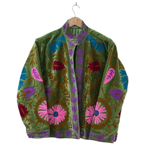 An olive green velvet bohemian jacket on a wooden hanger. It features traditional Suzani embroidery with large pink daisy-like flowers near the hem, bright blue and red leaves, winding bronze vines, and a contrasting purple embroidered border along the stand collar, front opening, and cuffs.
