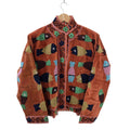 A rust orange velvet statement jacket on a wooden hanger. It features an eclectic all-over pattern of embroidered fish in various colors including dark blue, seafoam green, and mustard yellow. The stand collar, front opening, and cuffs are detailed with contrasting embroidered swirls and small green flowers.