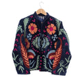 A dark navy blue velvet bohemian jacket on a wooden hanger. It features traditional Suzani embroidery with large orange and pink flowers on the chest, rust-colored paisley-like leaves on the sides, light green ferns, and pink chevron accents. The jacket has an open front revealing a striped inner lining.