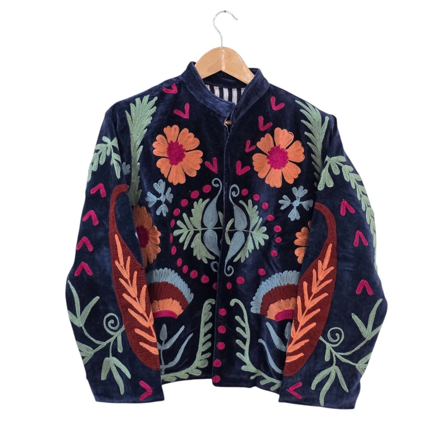 A dark navy blue velvet bohemian jacket on a wooden hanger. It features traditional Suzani embroidery with large orange and pink flowers on the chest, rust-colored paisley-like leaves on the sides, light green ferns, and pink chevron accents. The jacket has an open front revealing a striped inner lining.