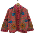 Front view of an open-style handmade coral pink textured cotton TNT jacket featuring vibrant bright blue, mustard gold, and green Suzani floral embroidery, showcasing an orange-and-white striped interior lining, hanging on a wooden hanger against a white wall.