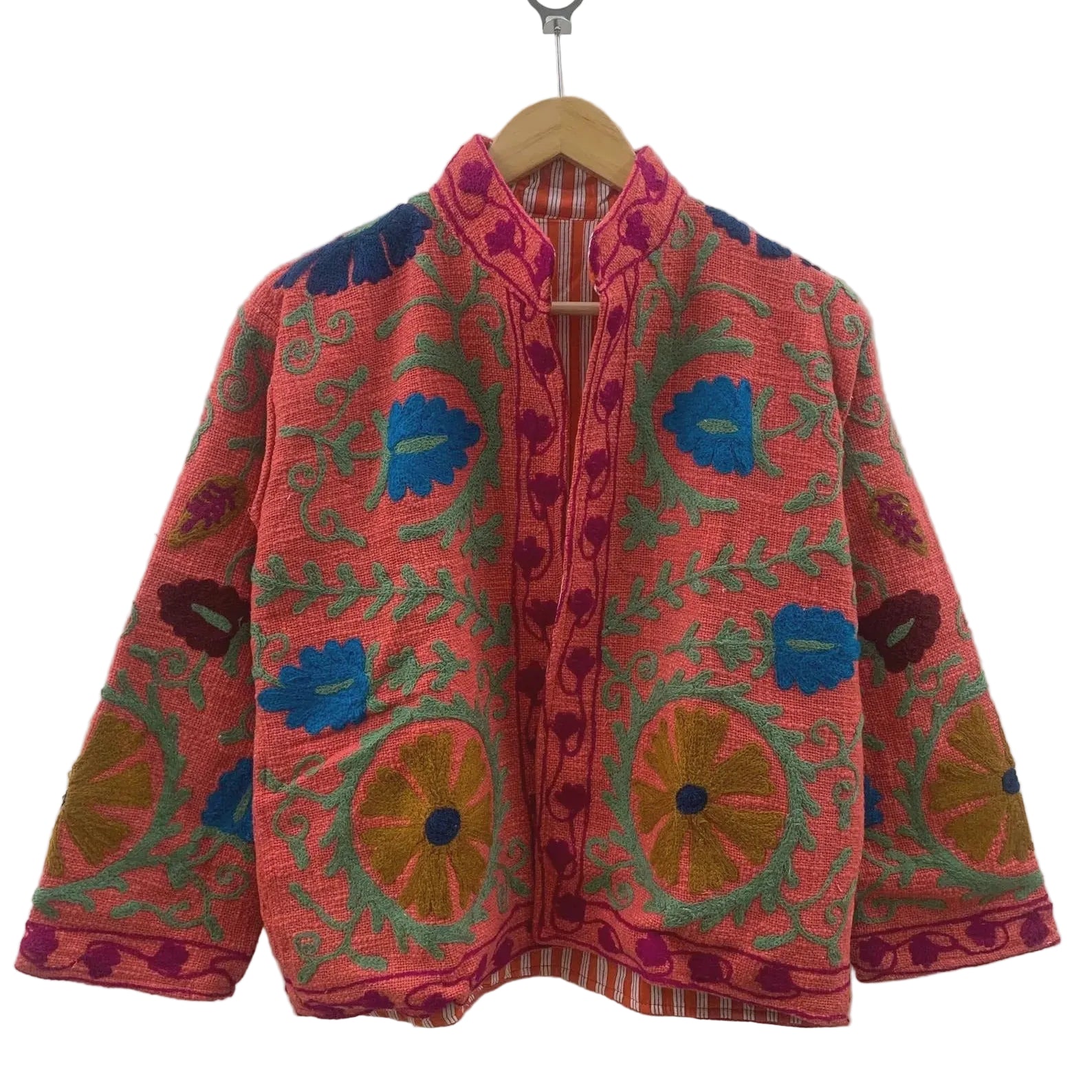 Front view of an open-style handmade coral pink textured cotton TNT jacket featuring vibrant bright blue, mustard gold, and green Suzani floral embroidery, showcasing an orange-and-white striped interior lining, hanging on a wooden hanger against a white wall.