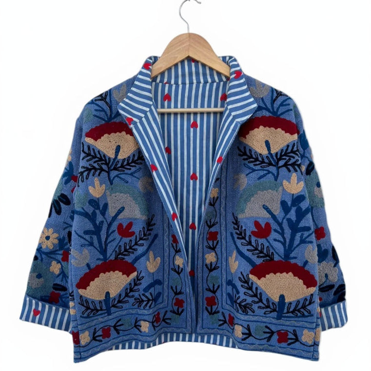 Front view of an open-style handmade denim-blue textured cotton TNT jacket with colorful Suzani floral embroidery, showcasing a blue-and-white striped interior lining dotted with small red hearts, isolated against a white background.