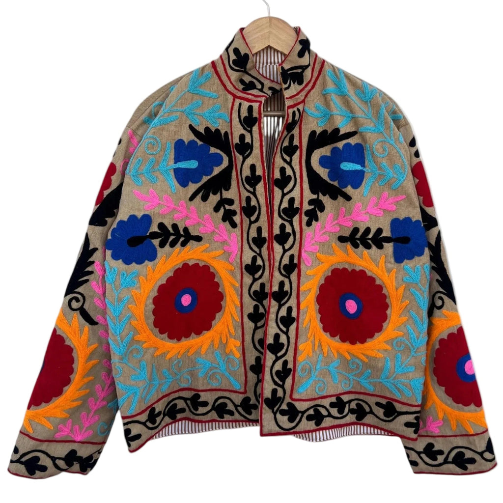 Front view of a handmade beige textured cotton TNT short jacket with bright multi-colored Suzani embroidery featuring large red and orange floral medallions with blue and pink accents, isolated against a solid white background.