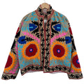 Front view of a handmade beige textured cotton TNT short jacket with bright multi-colored Suzani embroidery featuring large red and orange floral medallions with blue and pink accents, isolated against a solid white background.