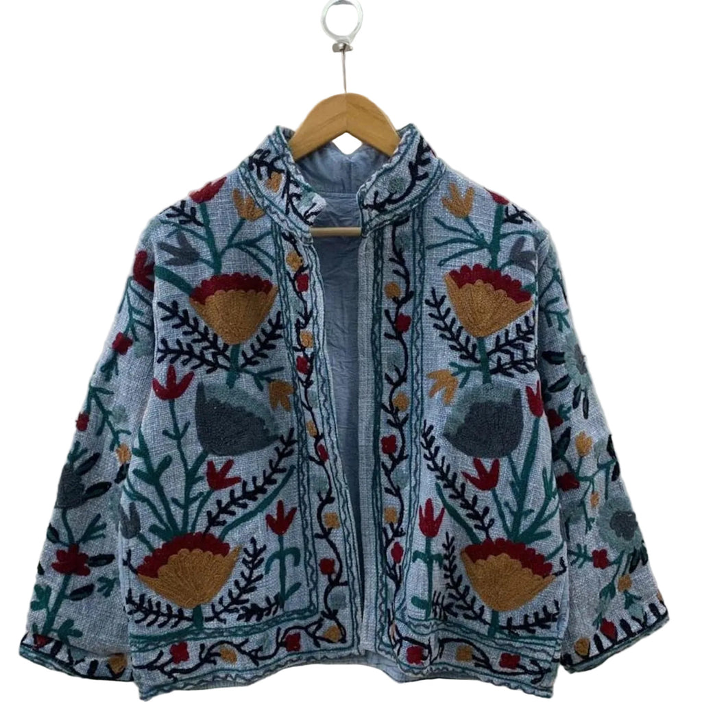 Front view of an open-style handmade slate blue textured cotton TNT short jacket featuring mustard yellow, red, and charcoal grey Suzani floral embroidery with dark green vines, showcasing a matching solid interior lining, hanging on a wooden hanger against a white wall .