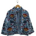 Front view of an open-style handmade slate blue textured cotton TNT short jacket featuring mustard yellow, red, and charcoal grey Suzani floral embroidery with dark green vines, showcasing a matching solid interior lining, hanging on a wooden hanger against a white wall .